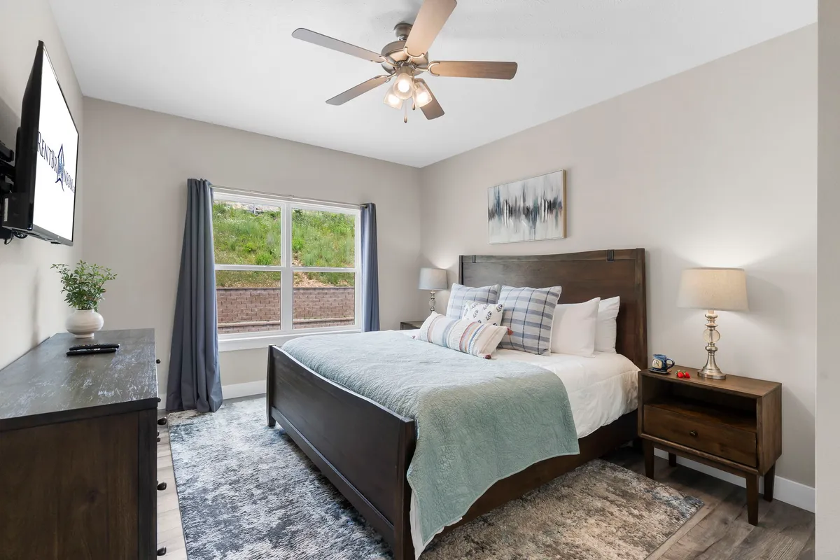 A cozy bedroom featuring a bed with decorative pillows, a nightstand with a lamp, a wall-mounted television, and a large window with curtains. The room exudes comfort, inviting guests to relax, read, or enjoy a movie night in bed while basking in natural morning light.
