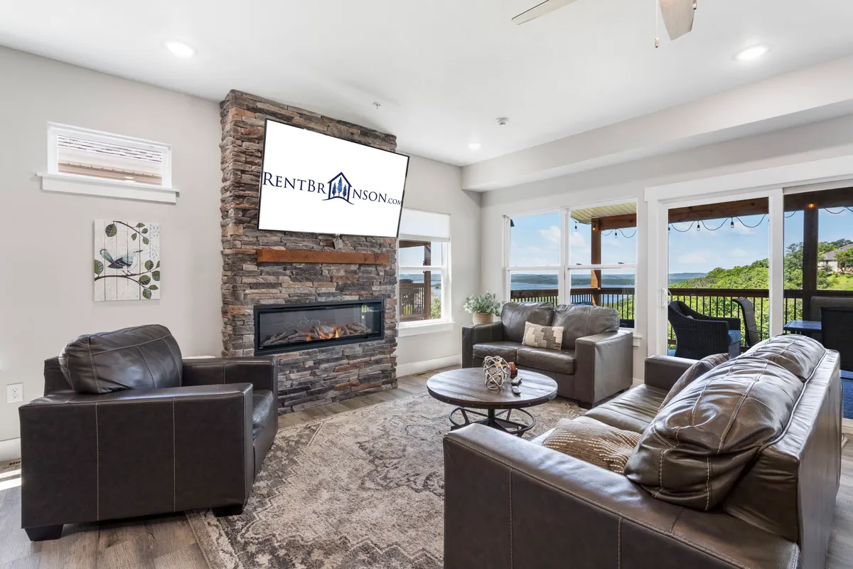 This image features a spacious living area with a stone fireplace, large television, plush seating, and expansive windows offering views outside. The décor is modern yet inviting, highlighting both relaxation and upscale finishes.