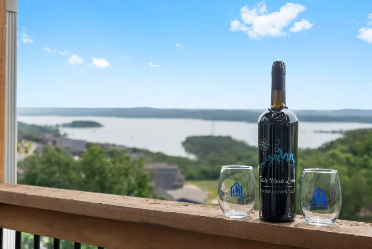 A stylish balcony setup with a wine bottle and two glasses perched on a railing, overlooking the sparkling expanse of Table Rock Lake. The scene exudes tranquility, with the calm water and distant shoreline visible in the background, suggesting the perfect spot for unwinding at sunset.