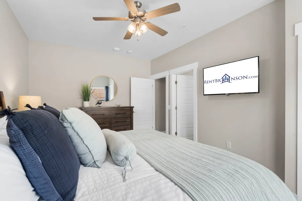 A cozy bedroom featuring a well-made bed with decorative pillows, a dresser, a circular mirror, and a television mounted for easy viewing from the bed. The space is inviting, designed for relaxation and private TV watching.