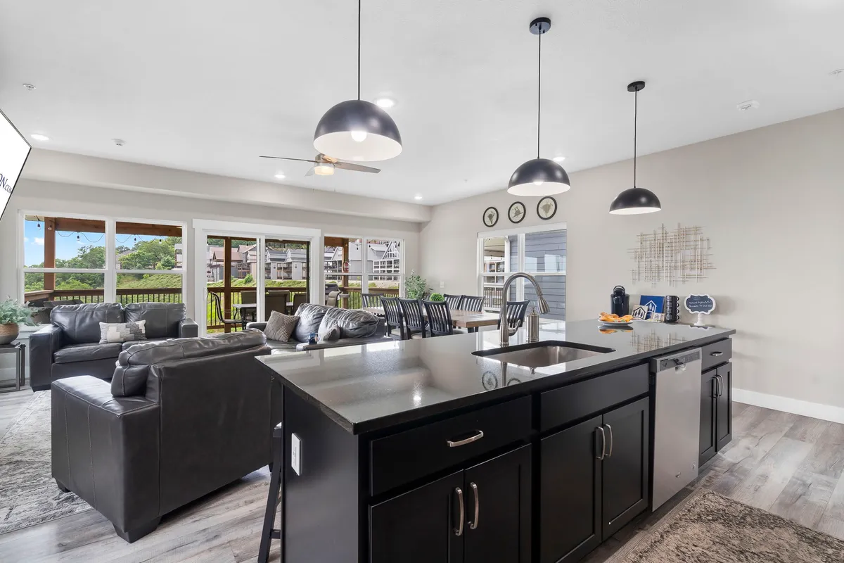 An open kitchen and living area with a kitchen island, sofa seating, dining area, and large windows. The design is modern, with high-end finishes and warm natural light, and the layout flows toward outdoor access.