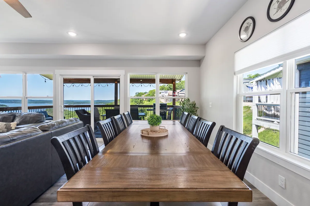 A welcoming dining area with a large table sits adjacent to windows framing the water, with comfortable chairs and a connected seating area. The table is set for gatherings, and sunset light spills in for an inviting feel.