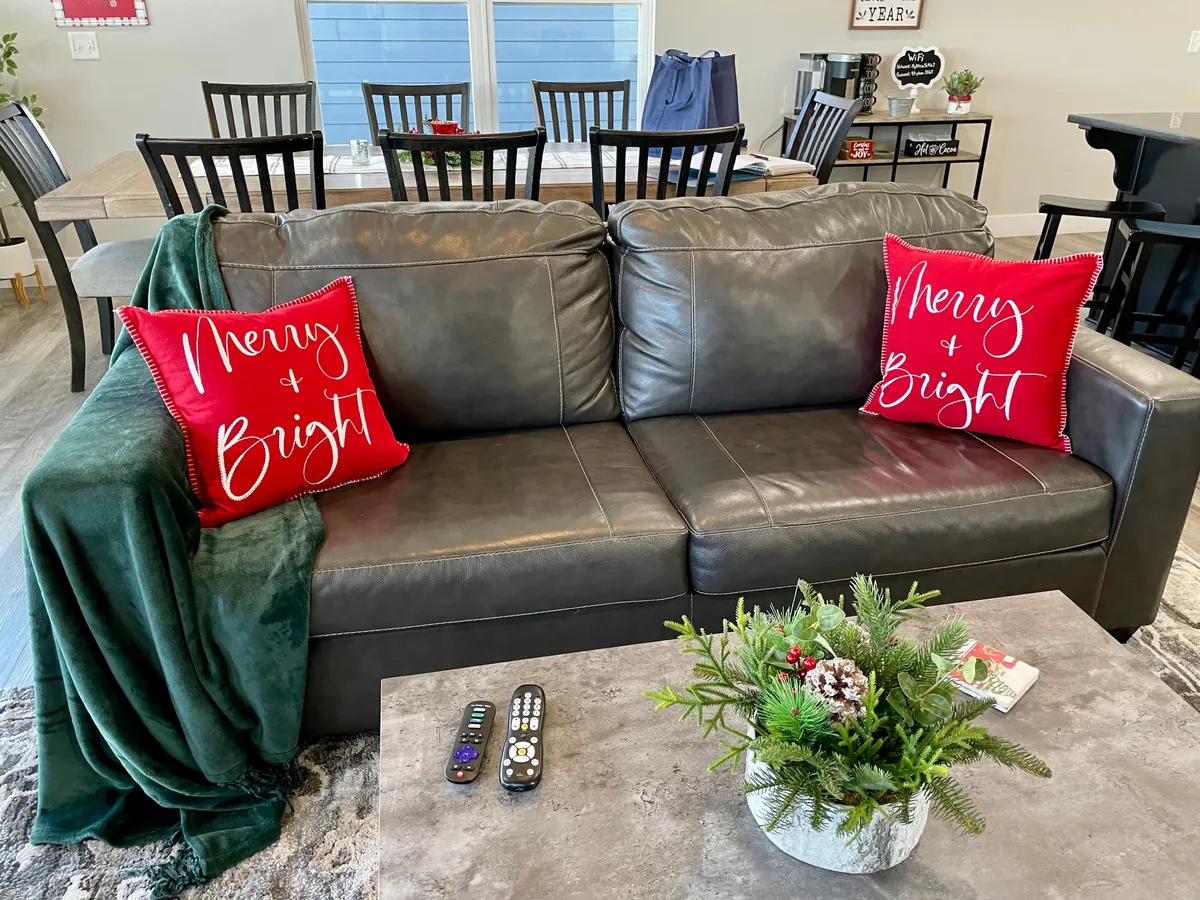 A spacious living area featuring a leather couch with decorative pillows, a coffee table adorned with greenery, and a dining space visible in the background. The setup is perfect for family gatherings, movie nights, and game sessions, with a warm and festive atmosphere.