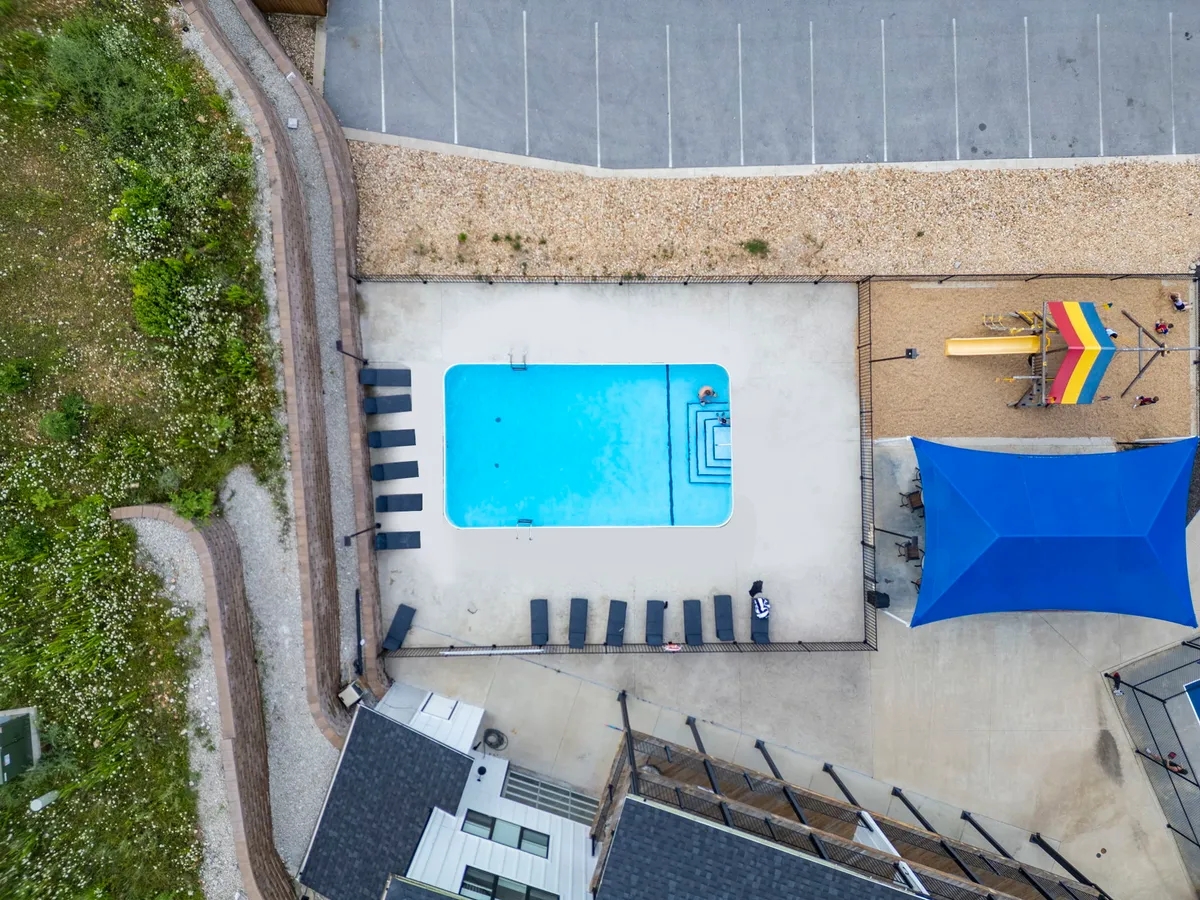 An inviting outdoor pool area with sun loungers and playground equipment nearby, beneath a shade structure. The space is bustling with leisure options for both adults and children, combining relaxation and play in a sunny, communal setting.