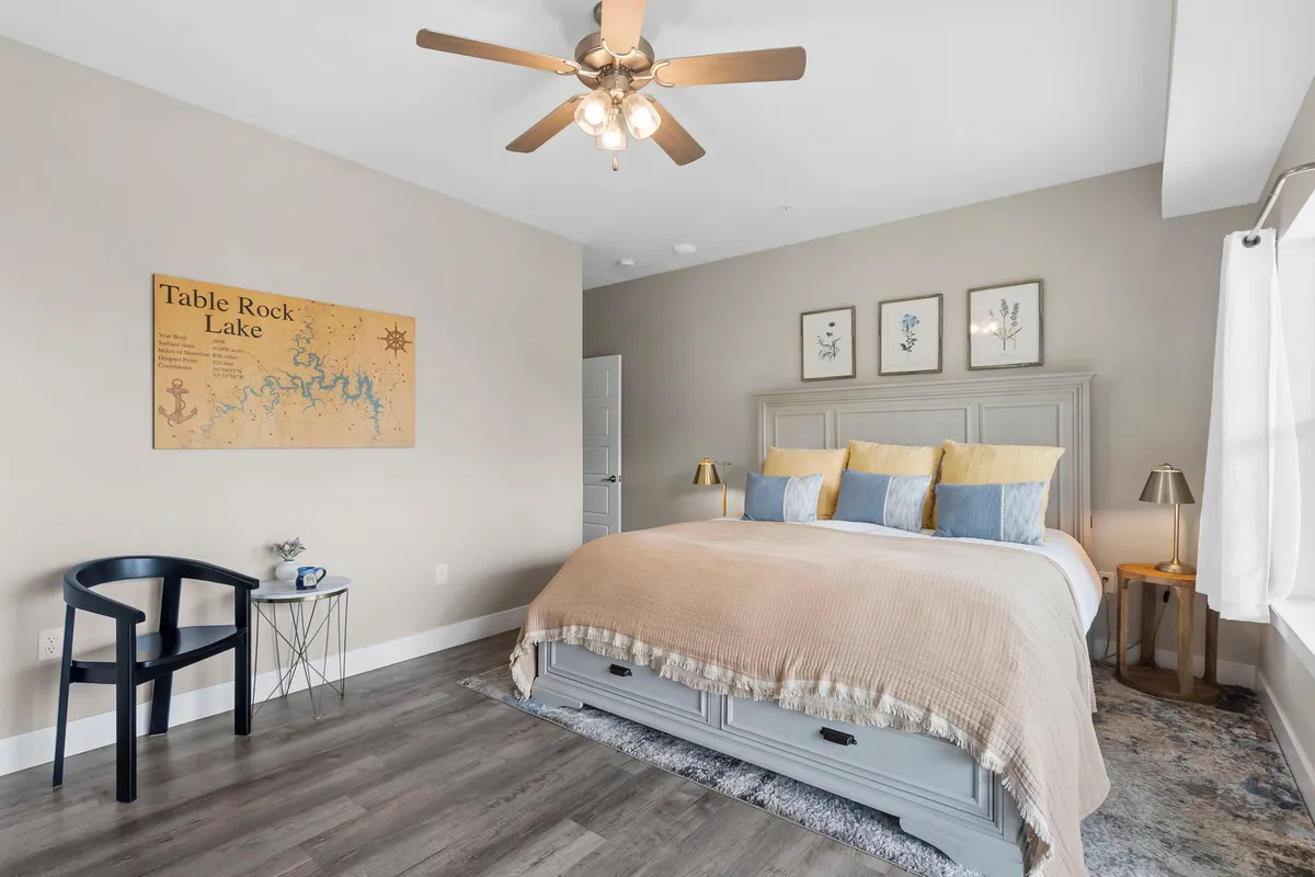 A tranquil bedroom with a plush bed adorned with decorative pillows, a map of Table Rock Lake on the wall, a comfortable chair and side table, and natural light streaming through the windows. The mood is peaceful and the space is designed for relaxing, reading, or enjoying a quiet cup of coffee.