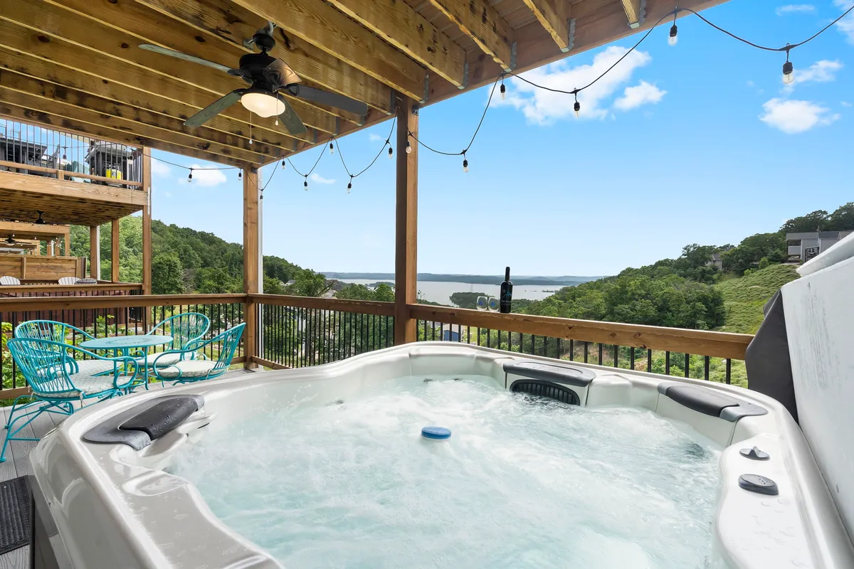 The image showcases the exterior of a stunning single-family home perched above Table Rock Lake, featuring a private hot tub overlooking sweeping lake views, an inviting outdoor dining set under rustic wooden beams, and a tranquil sunset atmosphere. The spacious patio is perfect for socializing or relaxing while taking in the breathtaking scenery.