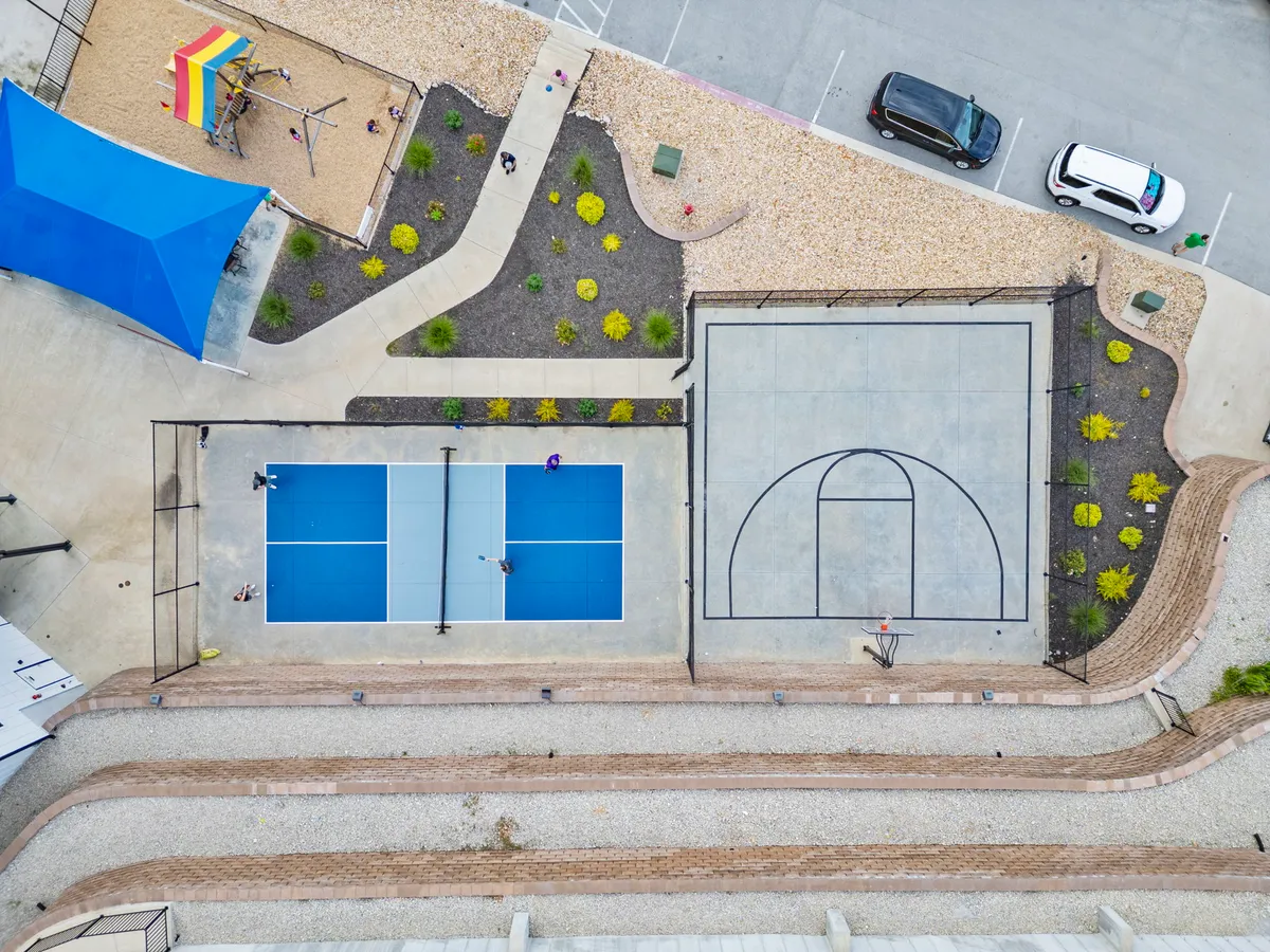 A vibrant exterior scene featuring tennis courts, a basketball court, and a colorful children's playground under a blue shade canopy. Landscaped areas and walking paths connect the amenities, creating a lively, family-friendly atmosphere.