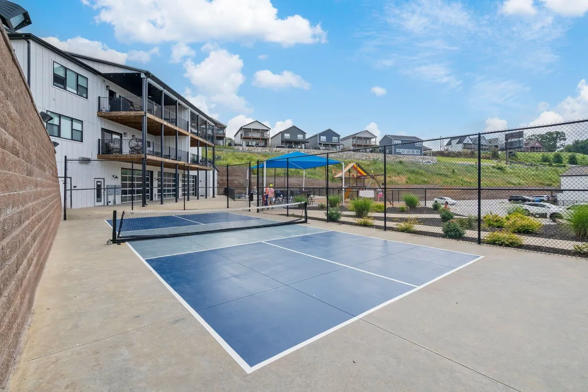 This exterior image zooms in on the tennis courts and playground, with residential buildings and lush landscaping forming a beautiful backdrop. The setting is active, showing people engaged in tennis and children playing, emphasizing social and recreational energy.