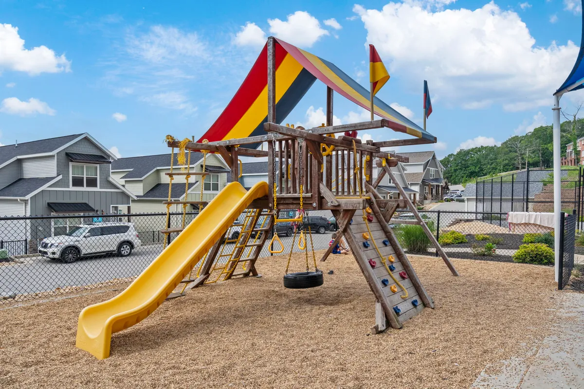 An exterior shot of a vibrant playground featuring a slide, tire swing, and colorful flags. Children are seen playing energetically while parents socialize nearby, creating a joyful and active community scene.
