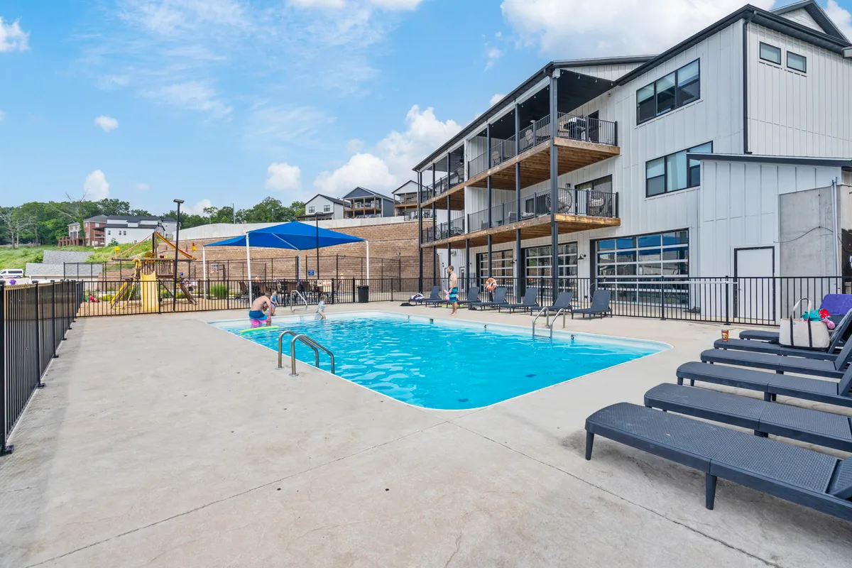 An exterior shot that captures the vibrant outdoor pool area, complete with lounge chairs and a visible playground in the background, while balconies from the building overlook the scene. Children are playing nearby, and the overall atmosphere is one of joyful summer fun and social connection.